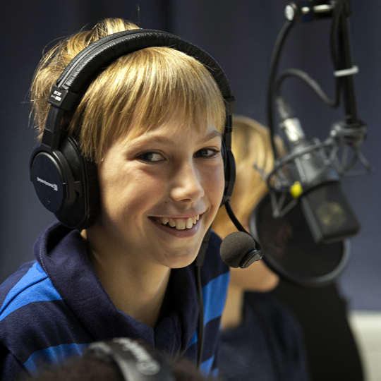 Junge mit Kopfhörern spricht lachend in ein Studiomikrofon beim Hörspiel-Kindergeburtstag
